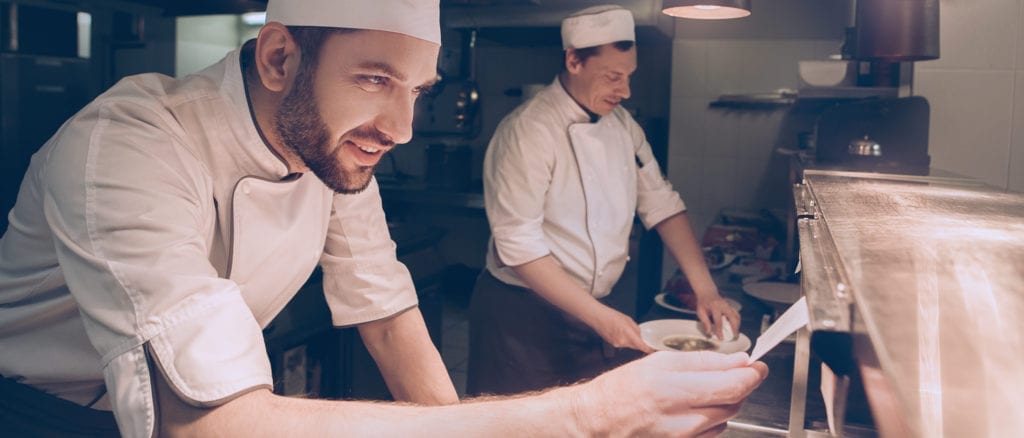 Smiling chef behind counter reviewing order ticket hanging from counter.